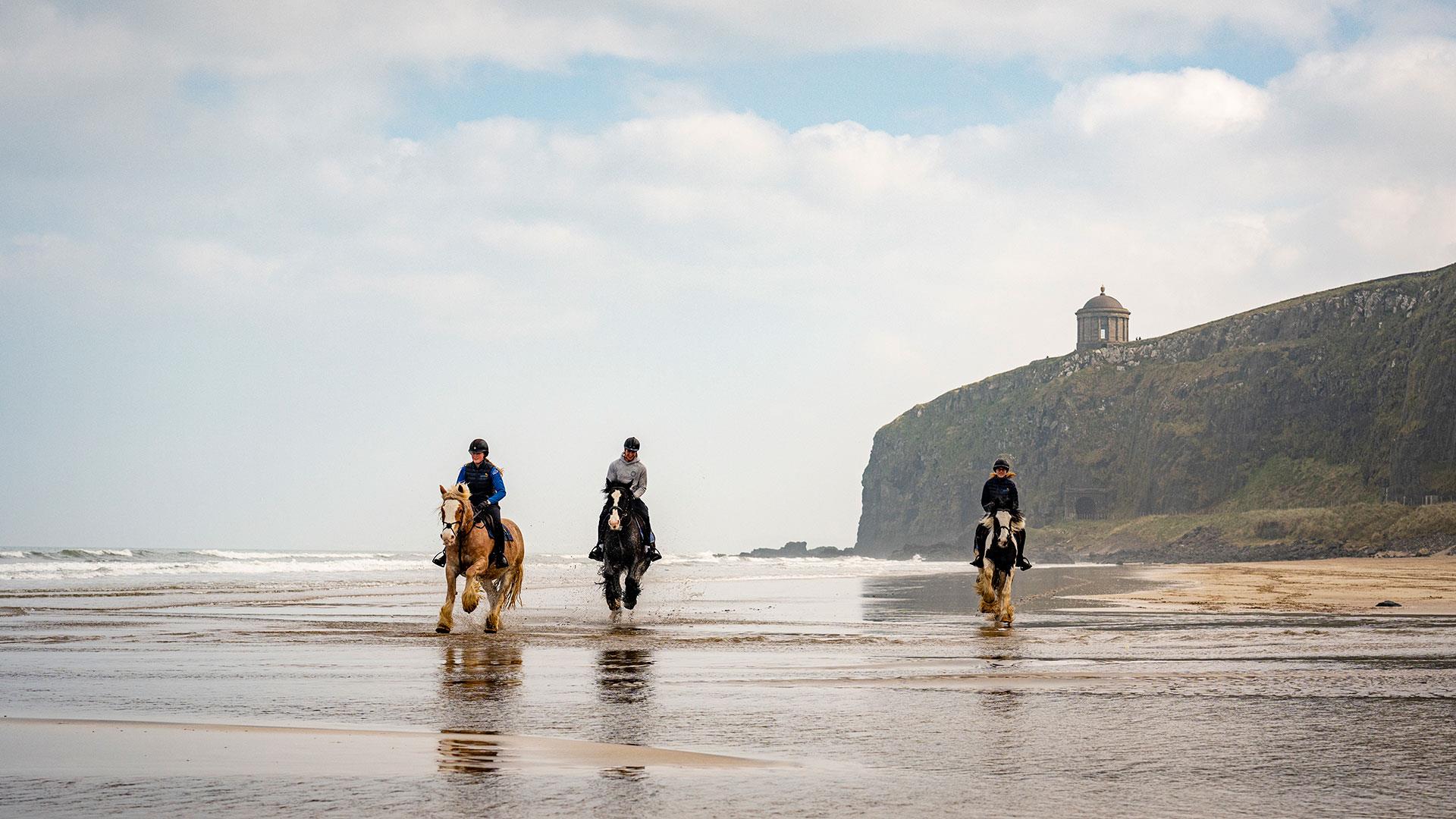 Group on horseback on the beach with Mussenden Temple in the background enjoying the Saddle Up by the Sea experience with Crindle Stables