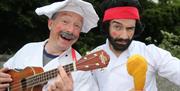 Two men dressed as chefs, one with a guitar,  one with a beard holding a rubber chicken leg