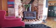 Image shows lounge area with comfy armchair, carpet and wood burner surrounded by lots of logs
