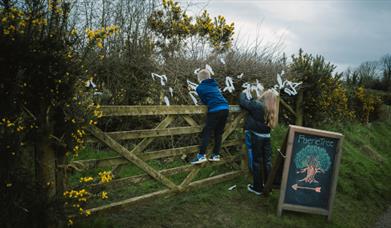 Children standing and climbing a country gate beside bushes with ribbons tied to them for May Day