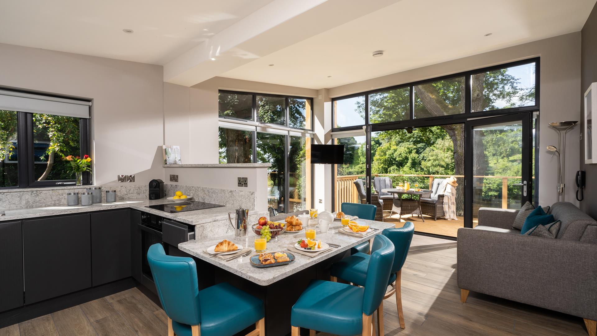 Open plan kitchen and dining area with view of lounge and outside area in background. Open patio doors floor to ceiling leading onto decked patio area