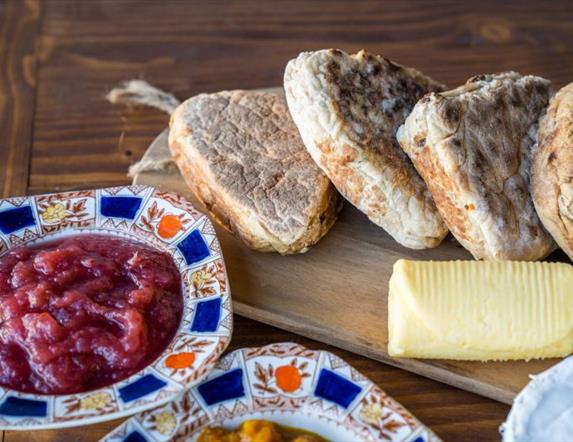 4 Soda breads with role of butter on a wooden bread board with jams in decorative bowls in the forefront