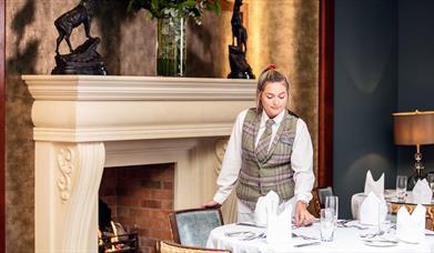 A Hastings Hotels member of staff setting a table in front of a fireplace.