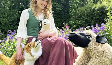 A female actress with long blonde curly hair sits with three puppets - a sheep, a fox, and a lamb on her lap.