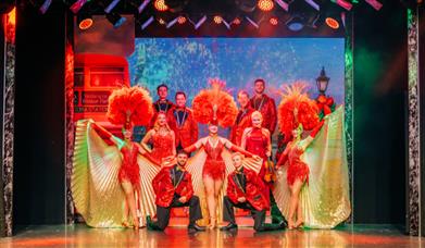 The cast showcases their talent on stage, surrounded by dramatic red feathers, creating a visually stunning spectacle.