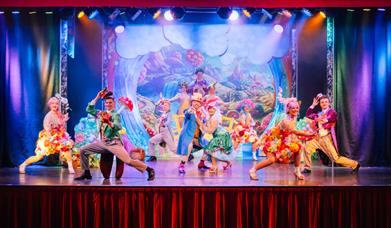 Performers in colorful costumes dance energetically on a stage with a vibrant, fantasy-themed backdrop.