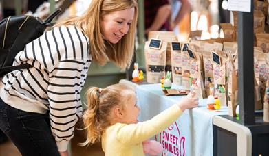 A little girl wearing a yellow top reaches for a bag of sweets. Her mum, who is wearing and white and black stiped top, crouches next to her smiling.