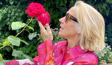 A woman in a colourful coat smells a red rose, celebrating nature and all things horticultural