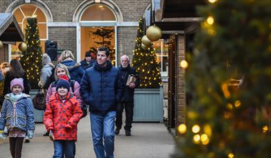 Two children and a man walk towards the camera wrapped in coats and scarves. People in the background are looking at stalls in wooden chalets. In the