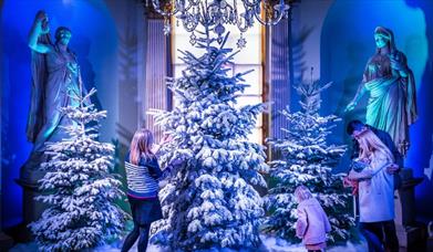 Three adults and a young girl look at three Christmas trees covered in fake snow which stand in Holkham's statue gallery, with roman statues looking o