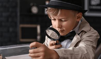 A boy in a black hat and bow tie looking through a magnifying glass.