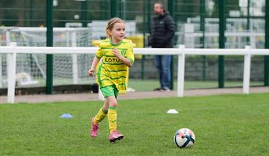 A young girl dressed in a yellow and green Norwich City Football Club kit runs towards a football, ready to kick it.