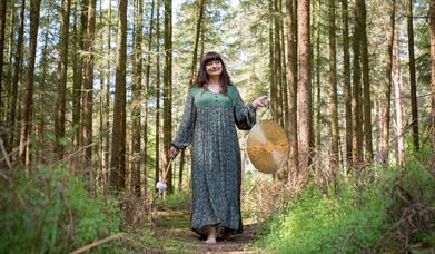 A white woman with long brown hair, wearing a green and brown patterned dress, walks though a forest holding a gong.