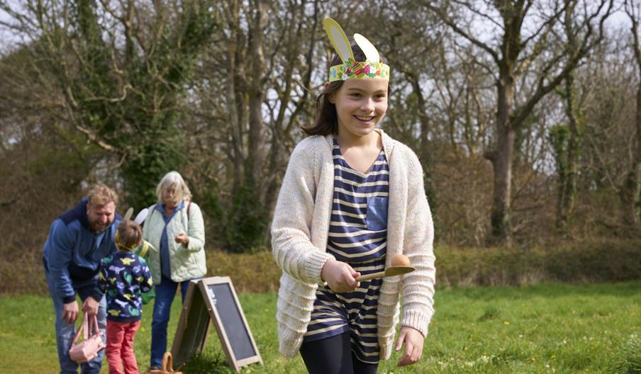 A girl balancing an egg on a spoon with family members in the background