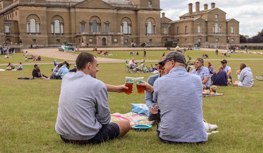 Two men cheers their compostable beers cups on a picnic blanket at outdoor food event in Holkham Park on the North Norfolk Coast
