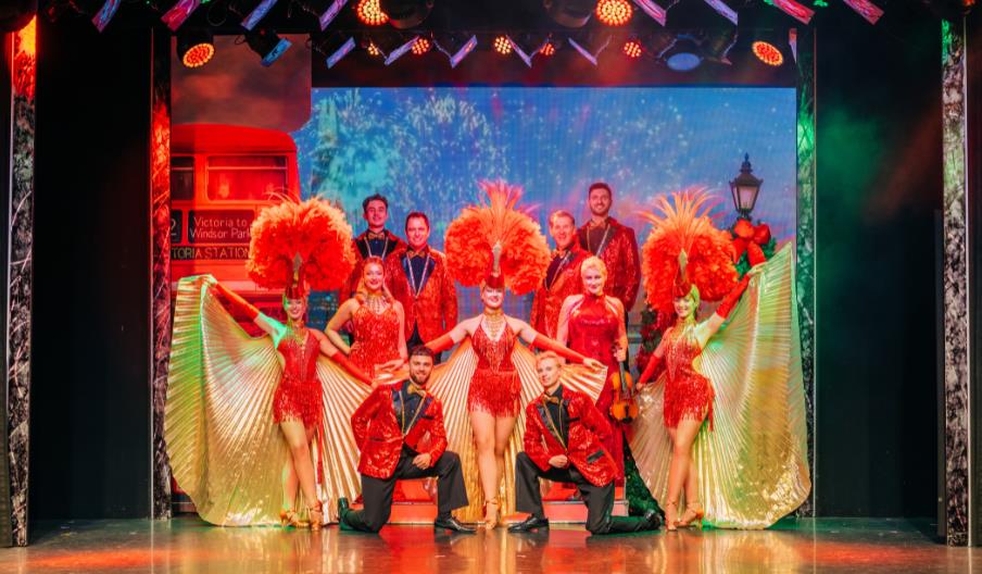 The cast showcases their talent on stage, surrounded by dramatic red feathers, creating a visually stunning spectacle.