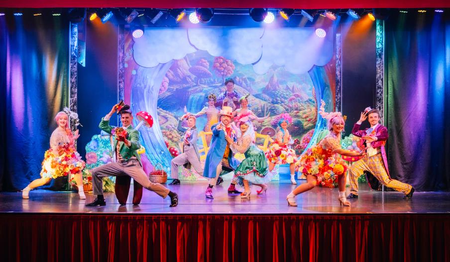 Performers in colorful costumes dance energetically on a stage with a vibrant, fantasy-themed backdrop.