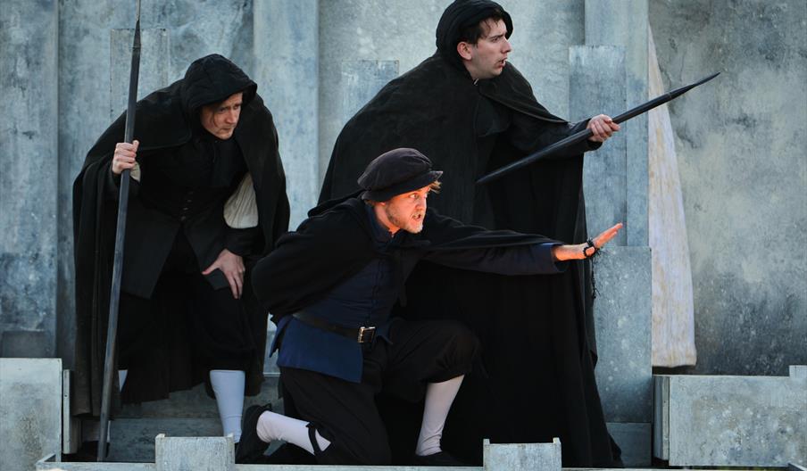 Three actors dressed in black ward off something out of shot, with concerned faces.