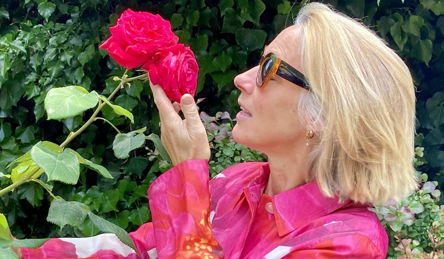 A woman in a colourful coat smells a red rose, celebrating nature and all things horticultural
