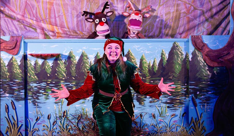 A performer dressed as a Christmas elf, wearing a red and green velvet costume with a Santa hat, stands smiling with open arms in front of a painted w