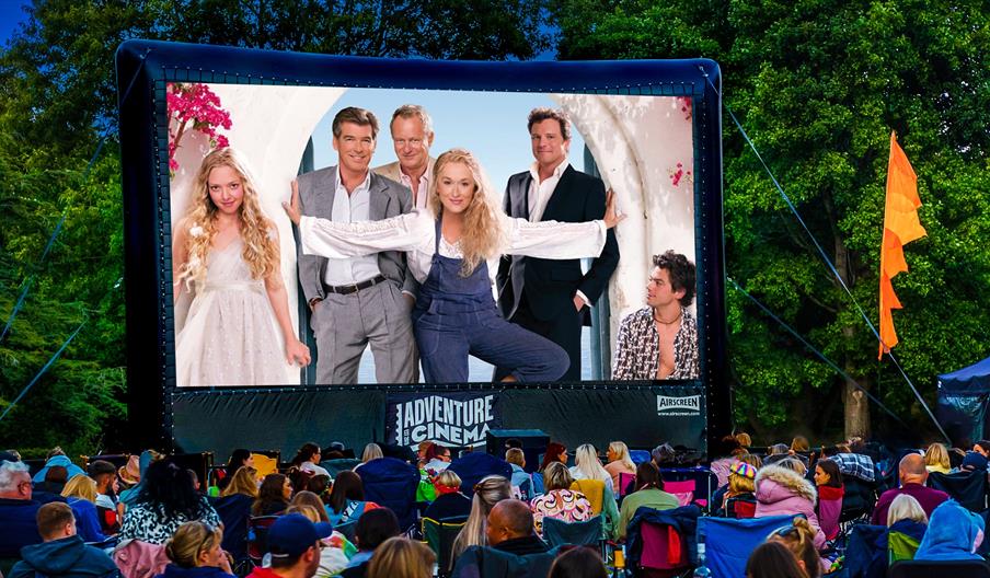 An outdoor cinema screen showing Mamma Mia! with lots of people sitting in deckchairs watching.