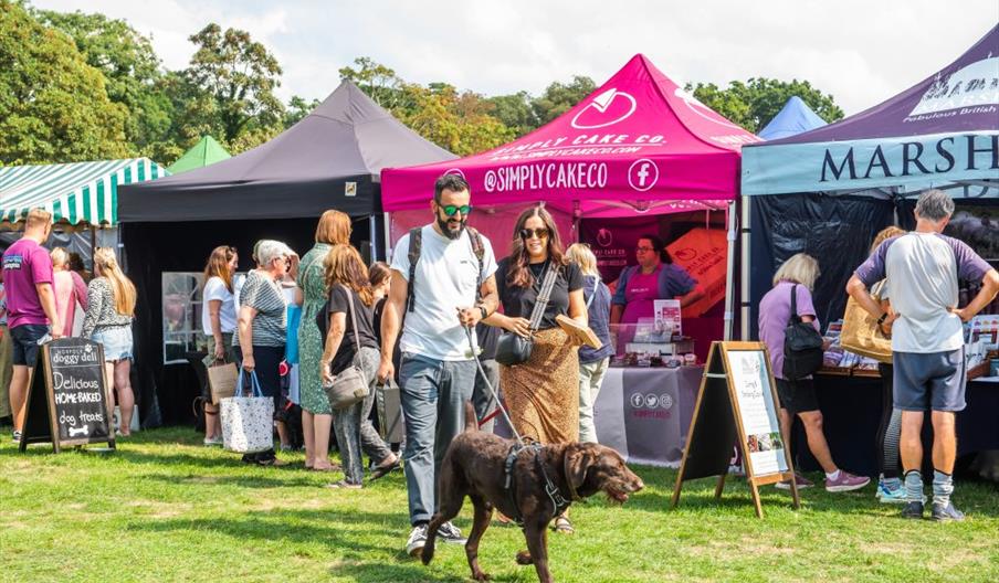 A couple wearing sunglasses walk towards the camera with their brown dog, holding a boxed purchase. In the background are market stall tents and gazeb