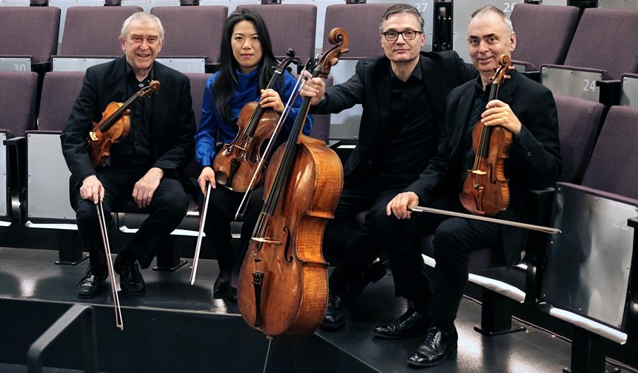 Four people holding string instruments while sitting on the seats of an empty venue.