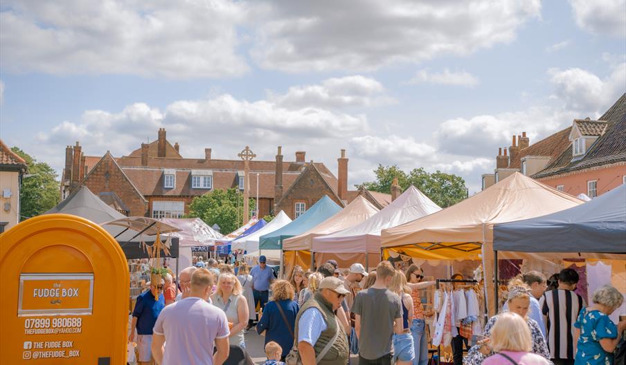 Crowds of people attending Holt Sunday Market. Gazebos line a closed road, creating a street carnival atmosphere on a sunny and bright day.