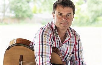A man casually holds a guitar while looking at the camera.