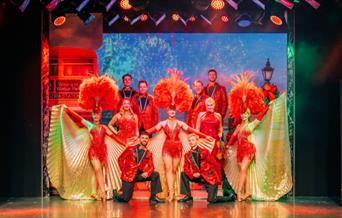 The cast showcases their talent on stage, surrounded by dramatic red feathers, creating a visually stunning spectacle.