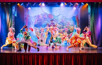 Performers in colorful costumes dance energetically on a stage with a vibrant, fantasy-themed backdrop.