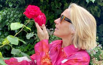 A woman in a colourful coat smells a red rose, celebrating nature and all things horticultural
