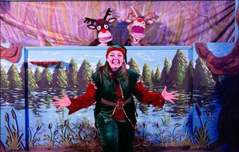 A performer dressed as a Christmas elf, wearing a red and green velvet costume with a Santa hat, stands smiling with open arms in front of a painted w