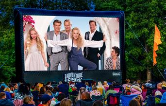 An outdoor cinema screen showing Mamma Mia! with lots of people sitting in deckchairs watching.