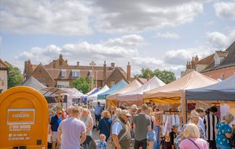 Crowds of people attending Holt Sunday Market. Gazebos line a closed road, creating a street carnival atmosphere on a sunny and bright day.