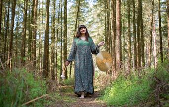 A white woman with long brown hair, wearing a green and brown patterned dress, walks though a forest holding a gong.