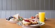 Three plates show a scone with cream and jam, a sandwich with salad, and a burger with salad. A tall glass of orange juice stands next to them.