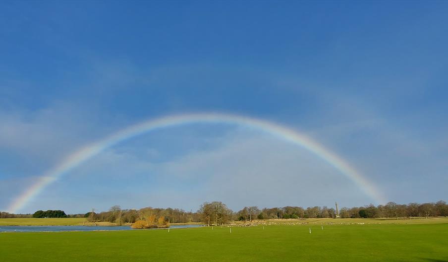 The image shows Holkham Park with the lake and monument. A rainbow appears over the Park.