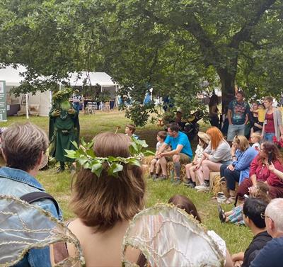A crowd sits in a circle on a field for the Sherwood Festival. They sit engaged in a story by a cosumted figure.