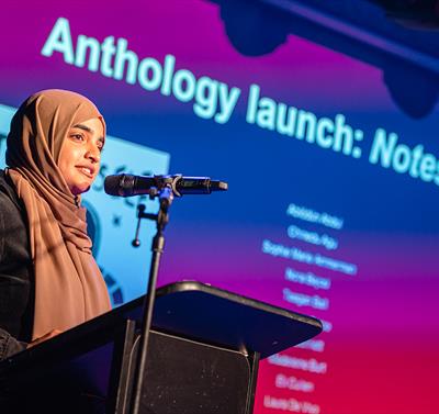 Image shows a girl on stage speaking into a microphone with a screen behind her reading - 'Anthology Launch: Notes to self.