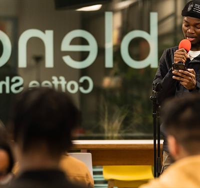 Image shows blend cafe sign in the background with a young boy holding an orange microphone wearing a black jacket, white top and black hat. There is