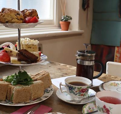 A serving of Afternoon Tea for two. A three tier cake stand showcasing sandwich squares, a mixture of cakes and fruit scones. Two teacups and saucers