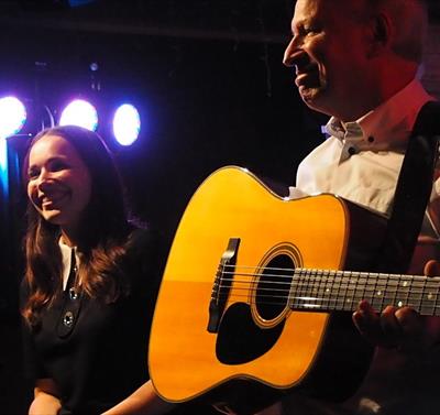 Image shows a man stood on stage playing a guitar and a woman stood next to him smiling.