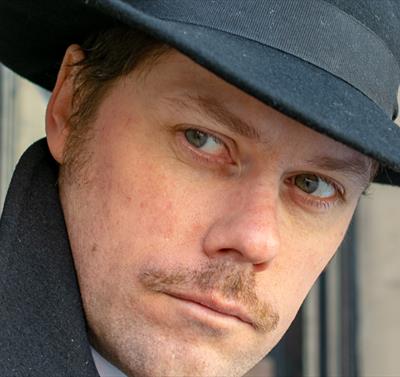 Man with fedora hat, pencil moustache and sinister appearance looks around a corner