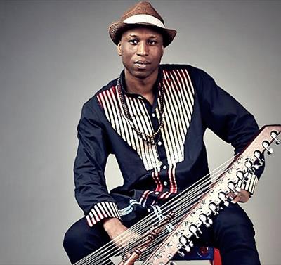 Image shows a man wearing a dark blue top with black and white stripes sat on a chair and holding an instrument.