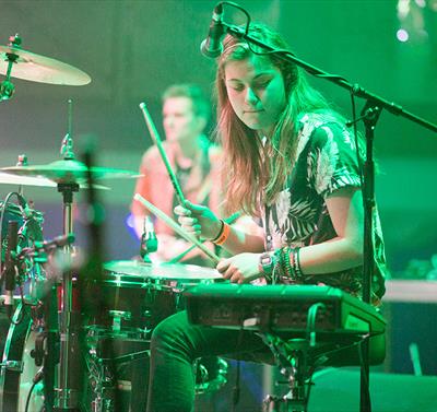 Image shows a girl on stage playing the drums with a microphone next to her.