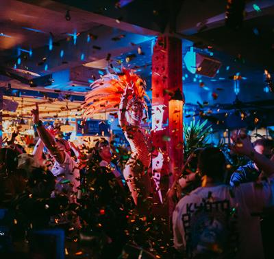 Photo of a crowded bar with fantastic dancing performers in feather headpieces, glitter faling from the ceiling and disco lights.