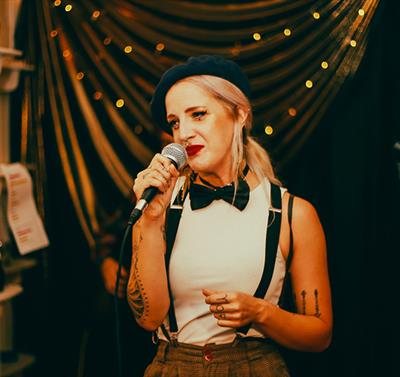 Image of young lady with blonde hair wearing a beret, bow tie and braces performing a spoken word piece on stage.