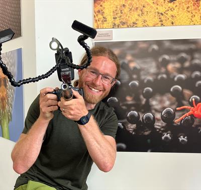 Alex Hyde holding a camera and smiling, with the sherwood forest up close exhibition wall behind