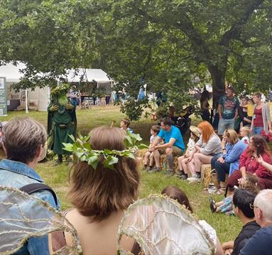 A crowd sits in a circle on a field for the Sherwood Festival. They sit engaged in a story by a cosumted figure.